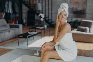 Women, cosmetology, beauty and hygiene concept. Relaxed thoughtful woman wrapped in bath towel, applies anti wrinkle cream or body lotion, poses in living room at home, looks pensively right
