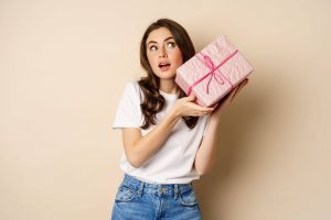 jpeg-optimizer_celebration-holidays-concept-happy-young-girl-looking-intrigued-shaking-box-with-gift-guessing-whats-inside-wrapped-present-standing-beige-background_wp